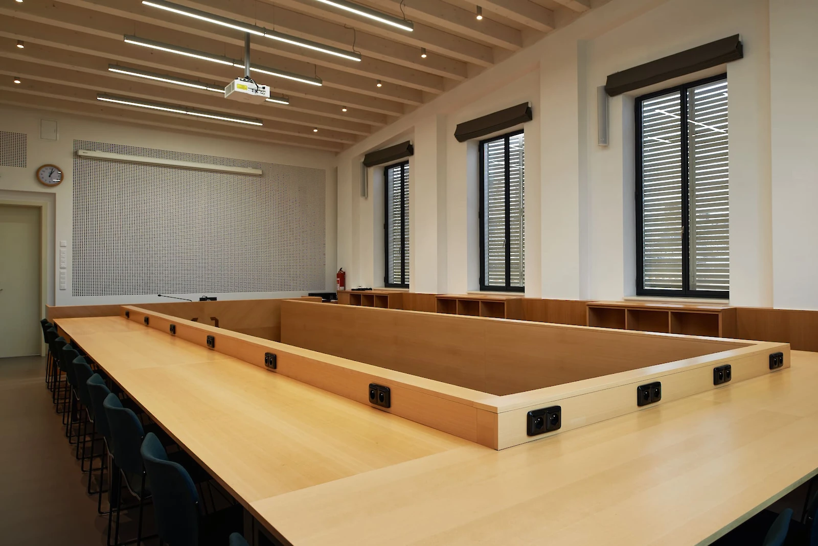 Učebna s dřevěným stolem kolem dokola se zásuvkami, židle okolo stolu